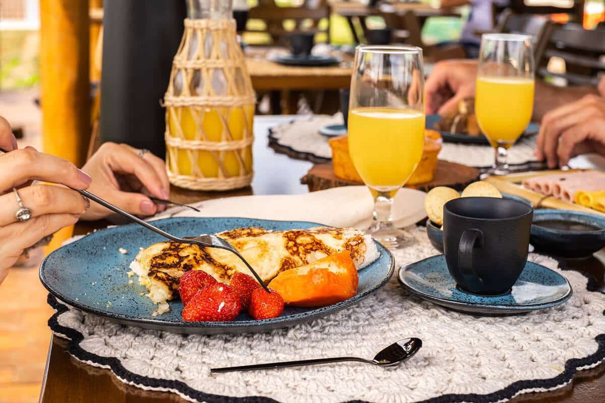 Prato de tapioca com morangos e mamão sendo servido na mesa do café da manhã, com suco de laranja, pão de queijo e frios ao fundo.