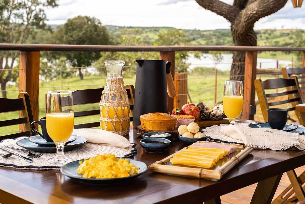 Mesa de café da manhã na varanda, com frutas, pães, bolo, suco e o lago ao fundo.