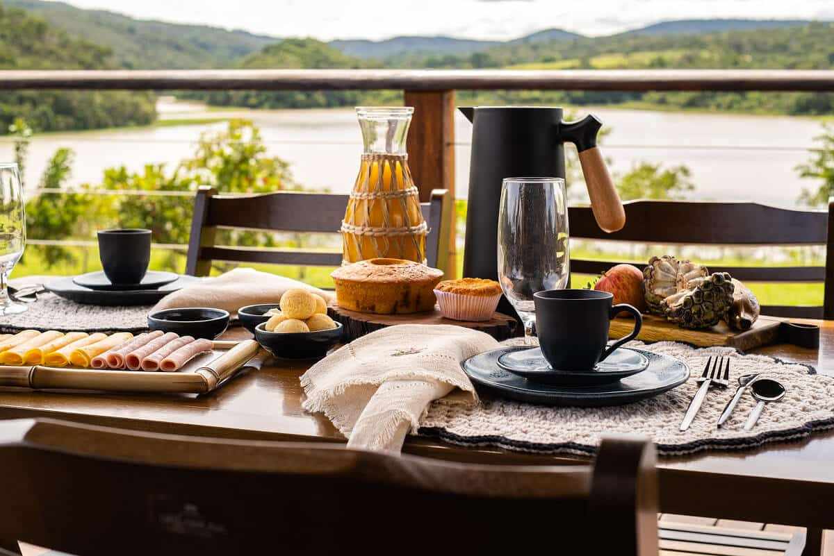 Detalhe do café da manhã com bolo, frios, garrafa térmica e o lago desfocado ao fundo.