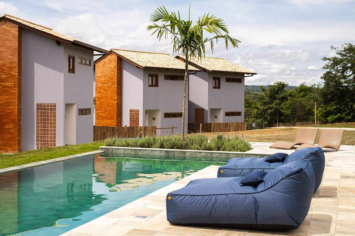 Fachadas dos chalés vistas da piscina, com palmeira ao centro e espreguiçadeiras azuis.