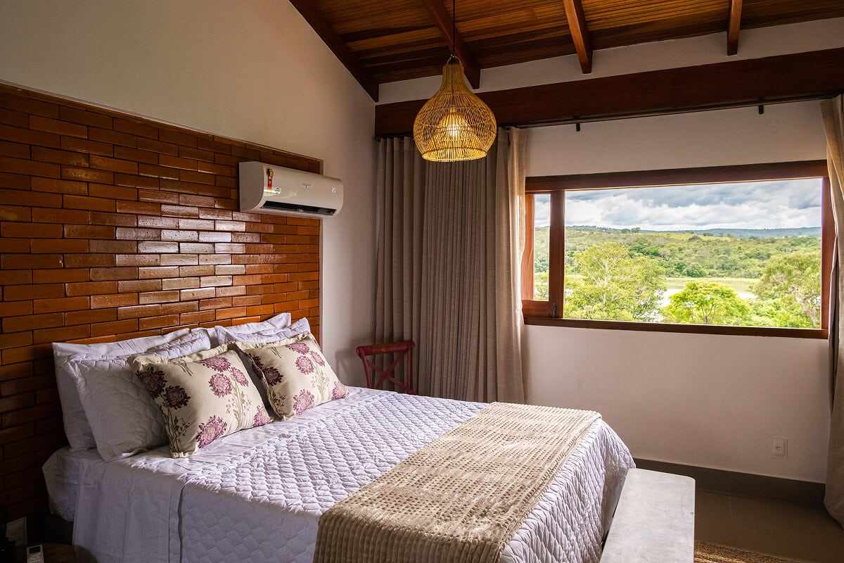 Quarto com cama queen, parede de tijolo, forro de madeira e vista para o cerrado e o lago.