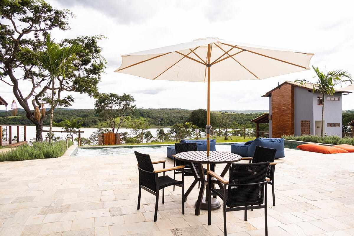 Mesa sob guarda-sol junto à piscina, com o lago e um chalé ao fundo.
