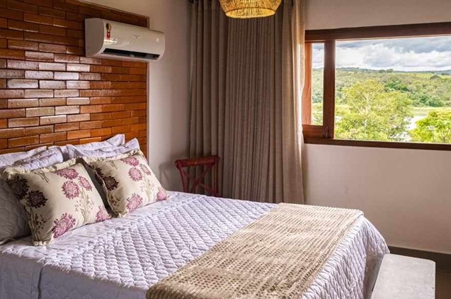 Cama queen com almofadas floridas e janela emoldurando a vegetação e a água.