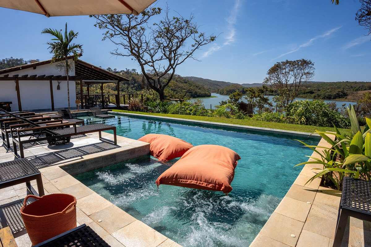 Piscina da Villa Sinhá Ana com almofadas flutuantes laranja, deck coberto à esquerda e o Lago Corumbá IV entre os morros ao fundo, em dia de céu azul.