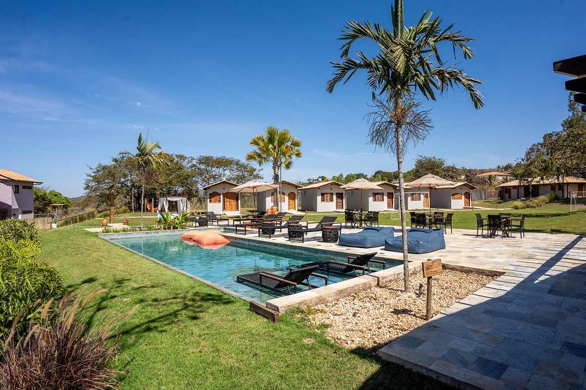 Piscina retangular com espreguiçadeiras dentro da água e a fileira de chalés de tijolo e reboco claro ao fundo, sob céu azul.