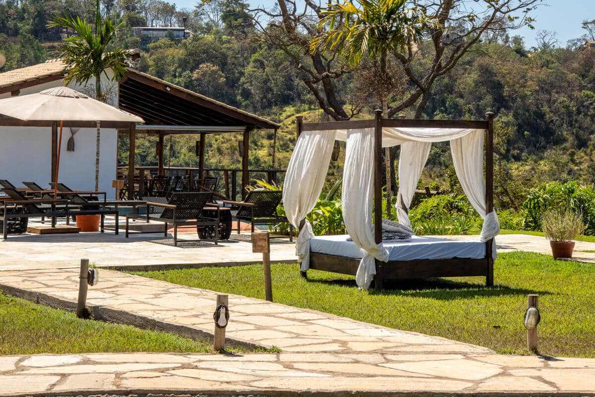 Cama balinesa com cortinas brancas montada no gramado, ao lado do deck coberto e da piscina, com a mata do cerrado atrás.