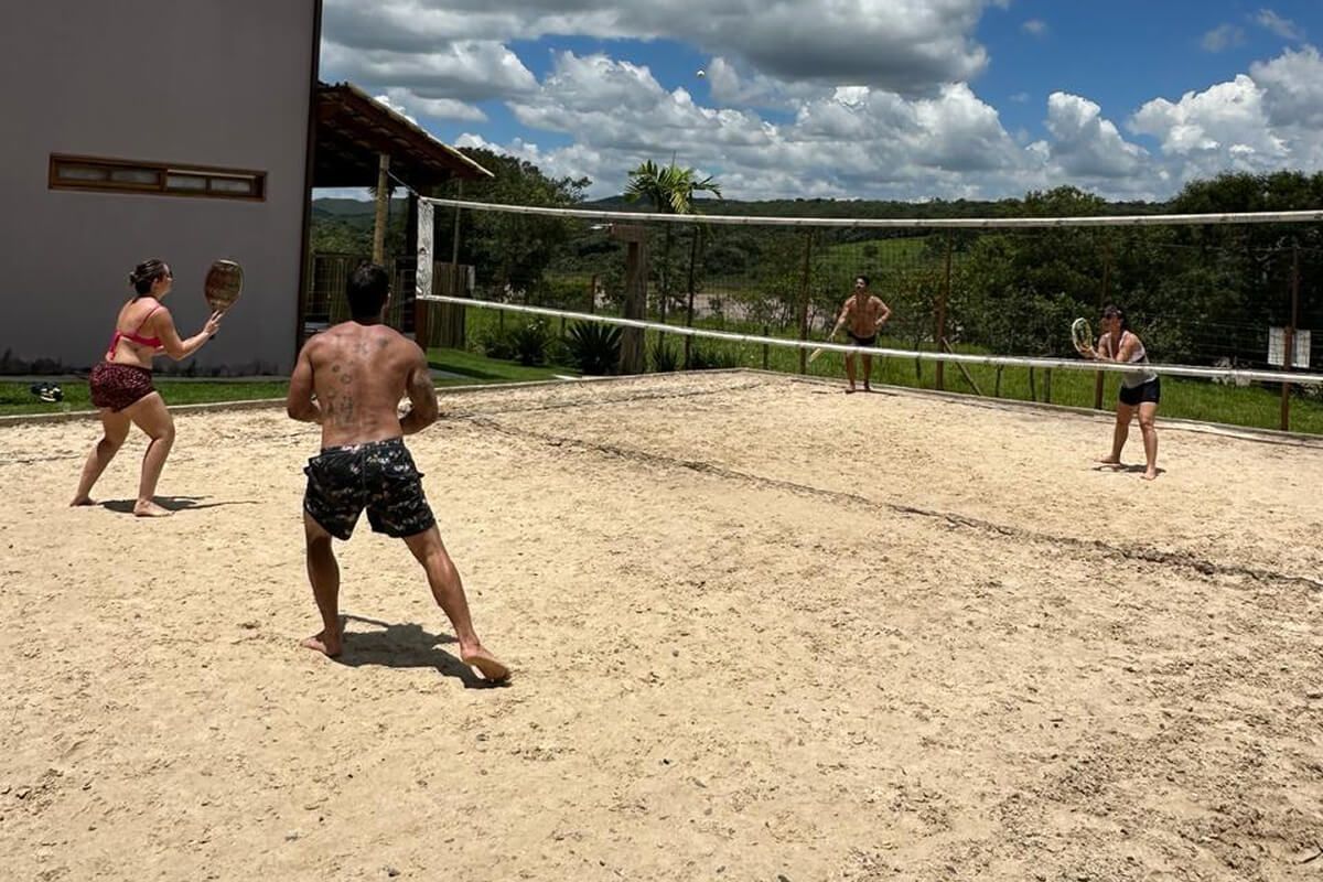 Quatro pessoas jogando beach tennis na quadra de areia da Villa Sinhá Ana, com rede armada, o cerrado e o céu de nuvens ao fundo.